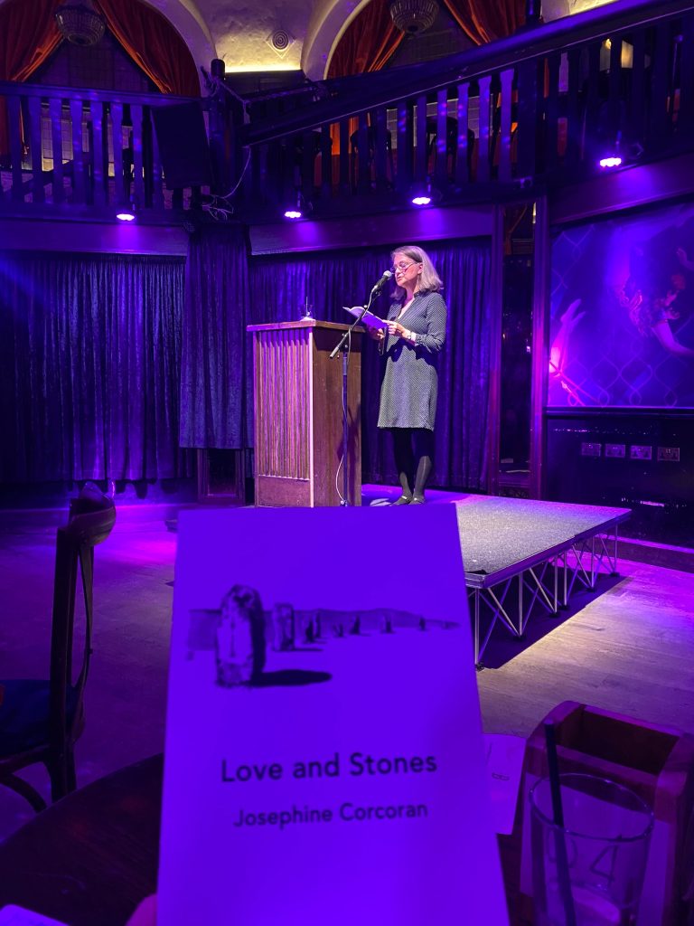 Josephine Corcoran reading on stage at a lowlit venue. In the foreground a picture of her book Love and Stones