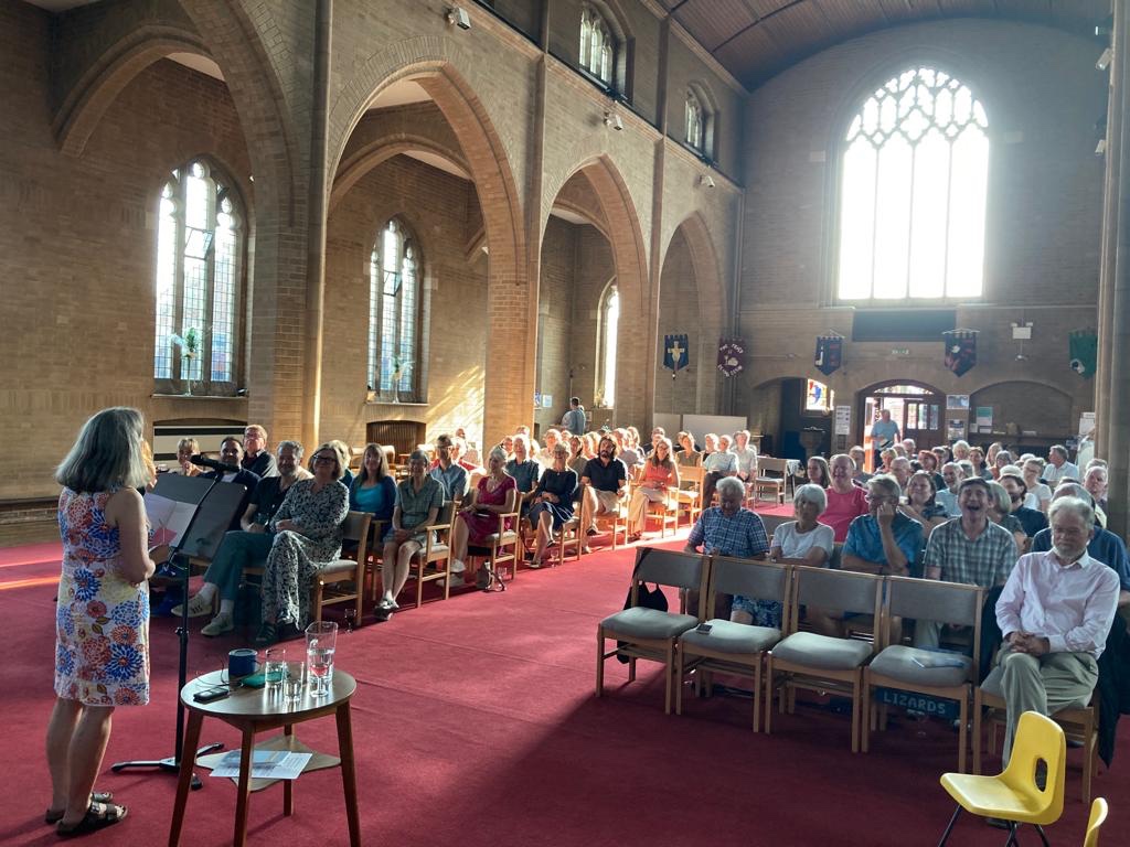 Taken from behind the reader and showing Josephine Corcoran reading to a large audience inside a church.