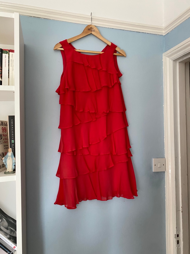 A sleeveless red silk dress with tiered frills of fabric, hanging on a coat hanger against a blue wall.