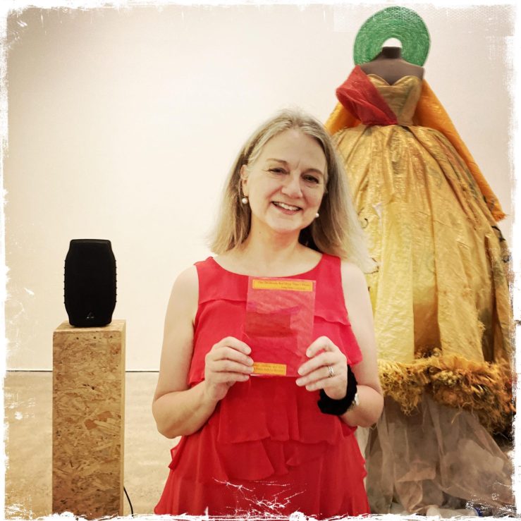 Josephine Corcoran wearing a red dress and holding her red-covered pamphlet, standing to the side of Sandra Suubi's artwork 'Samba Gown'.